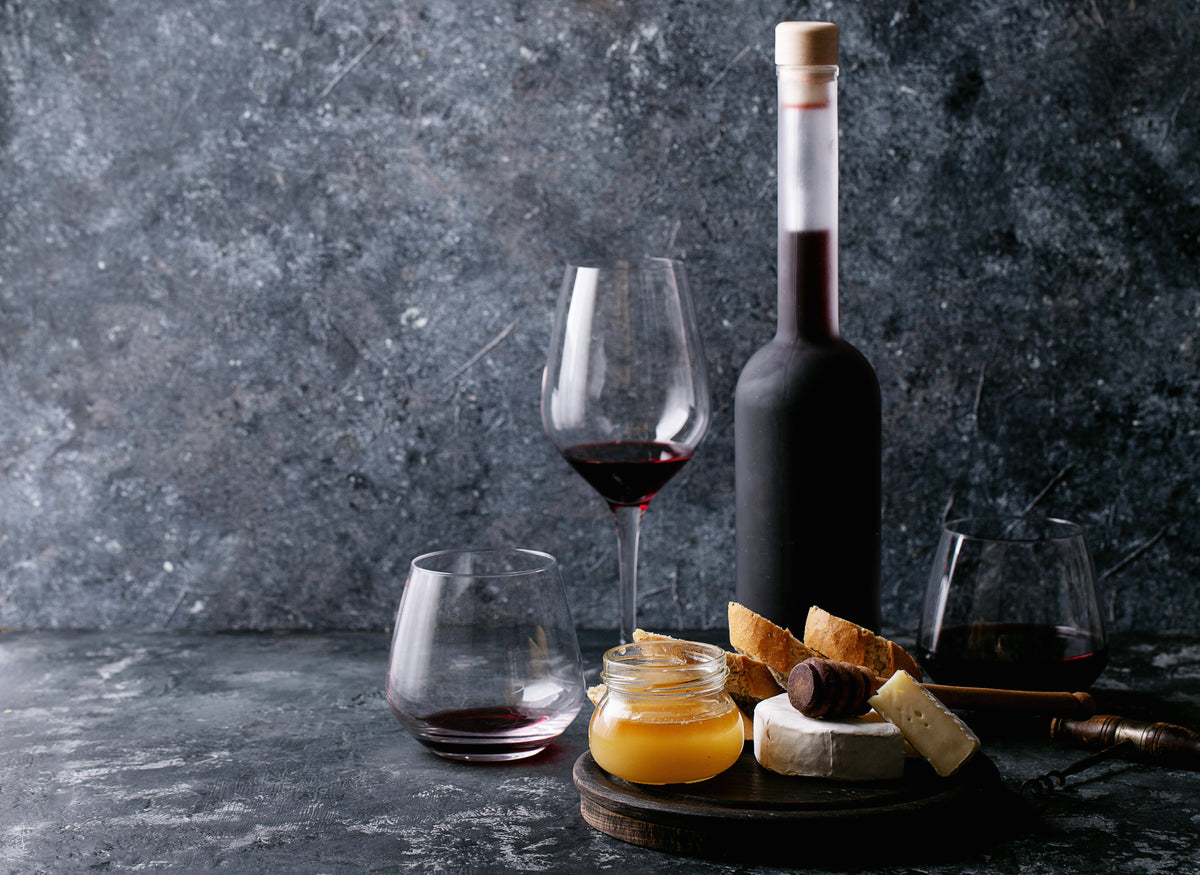 Wine bottle and glasses with cheese, honey and bread against a dark textured background.