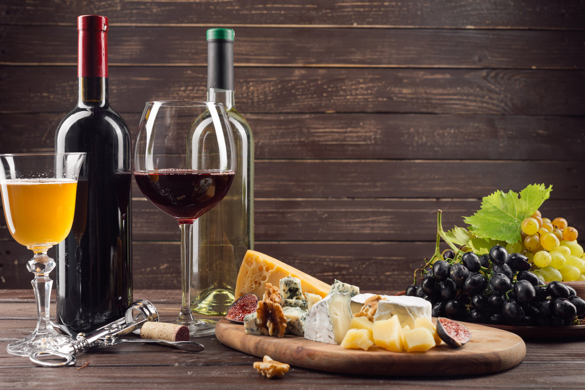 Wine bottles and glasses beside a cheese board, grapes, figs and walnuts on a wooden table.